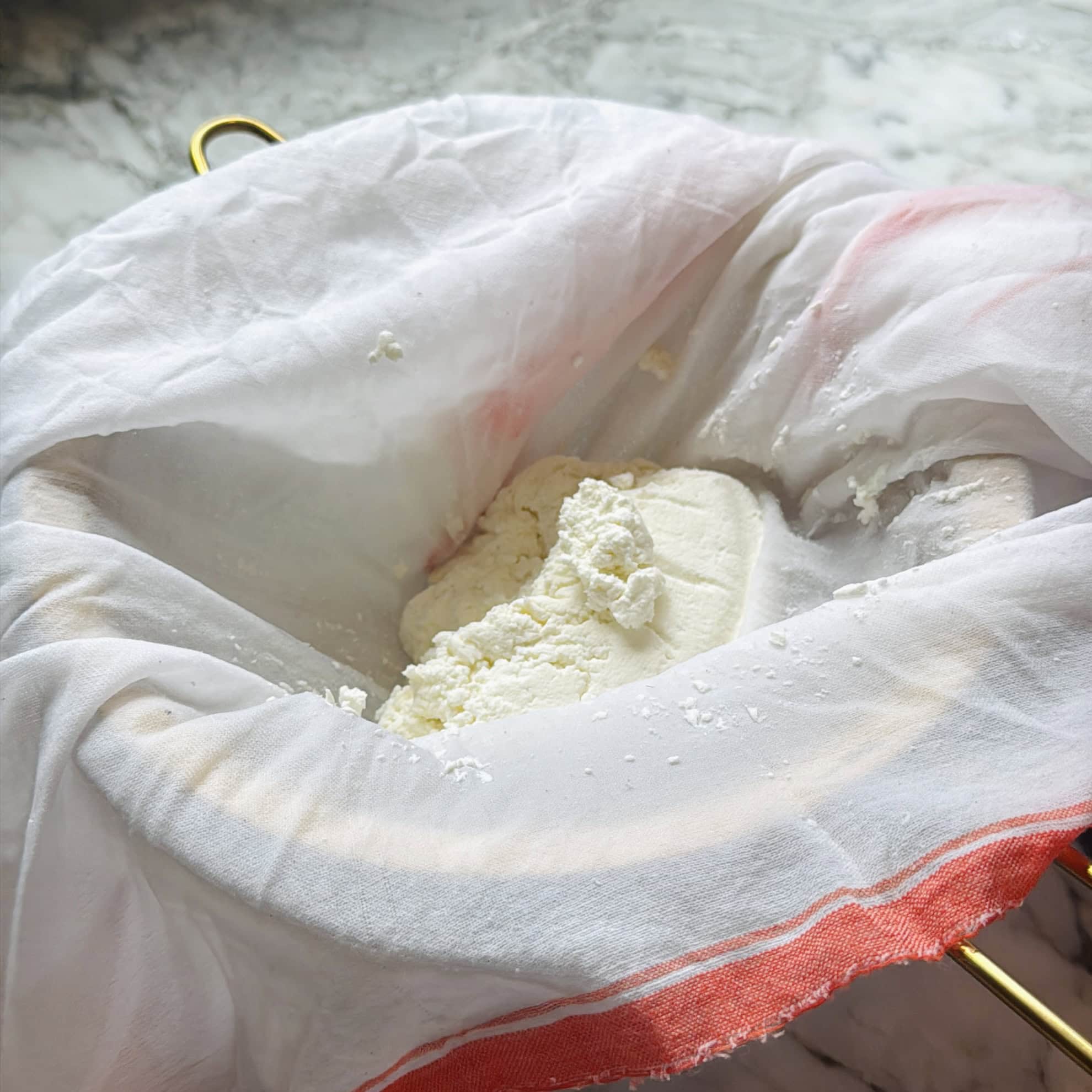 A cheesecloth-lined strainer with white cheese curds resting inside, placed on a marble countertop.