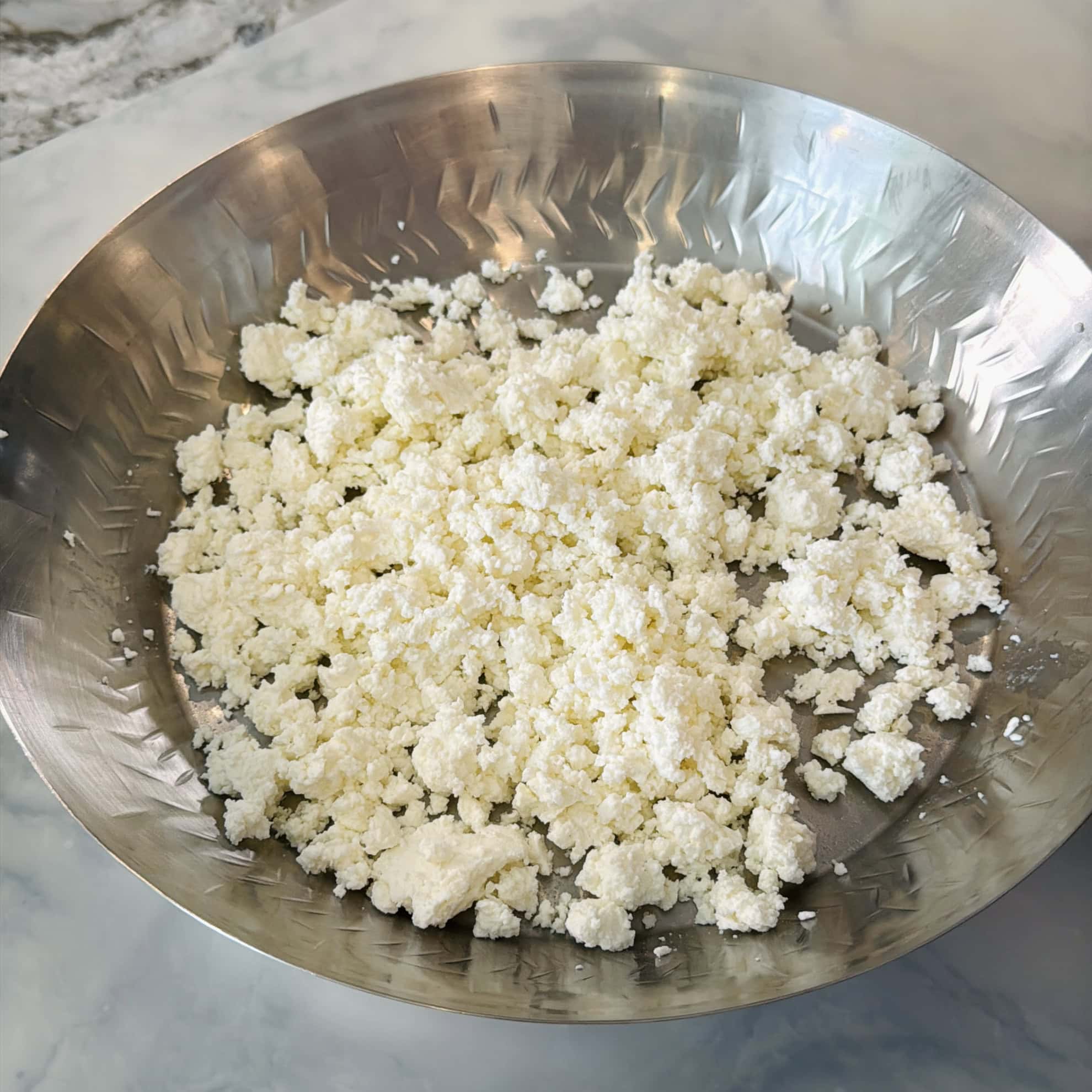 Crumbled white cheese is spread on a round, metal plate placed on a marble surface.