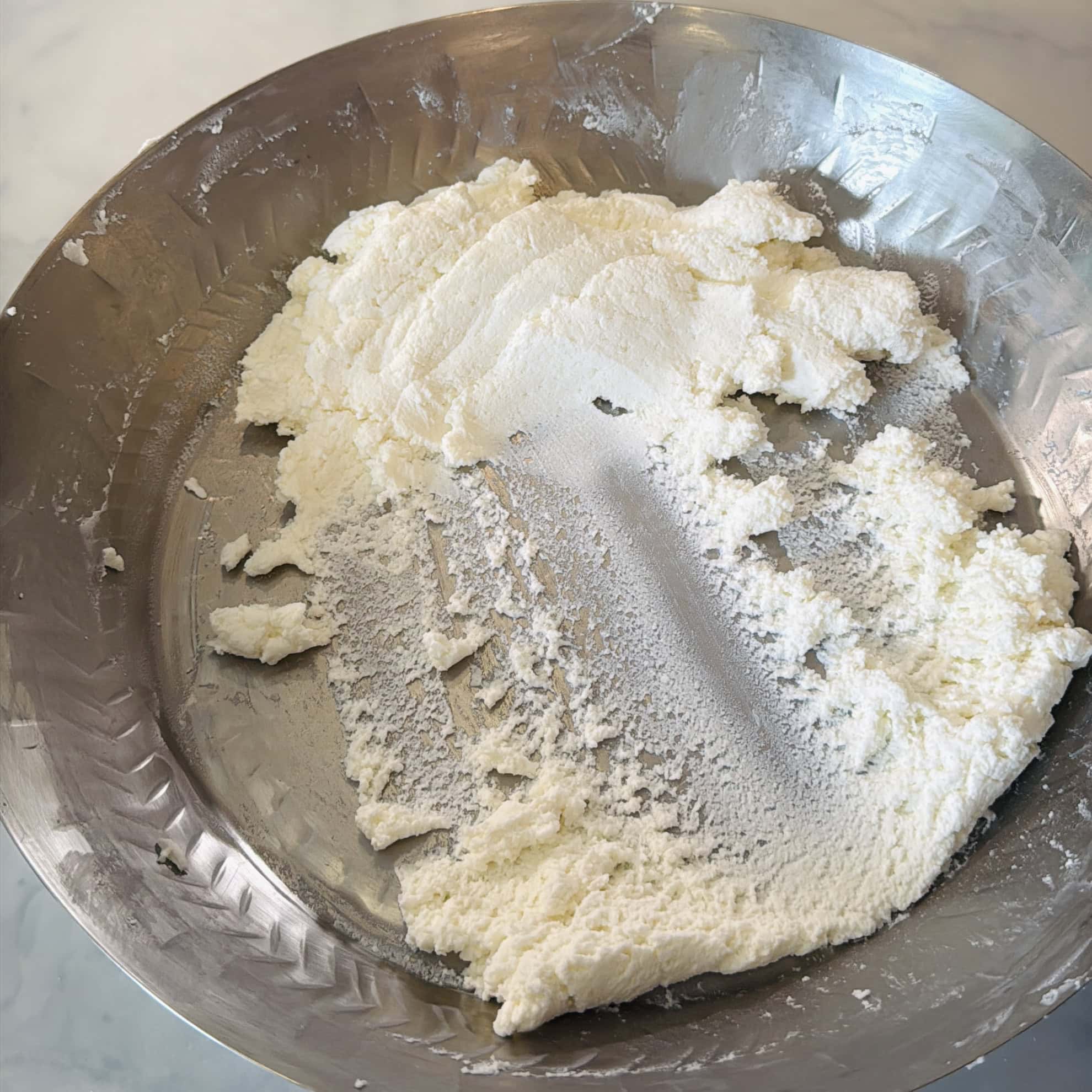 A shallow metal plate containing a partially spread layer of fresh, crumbly white cheese on a marble surface.