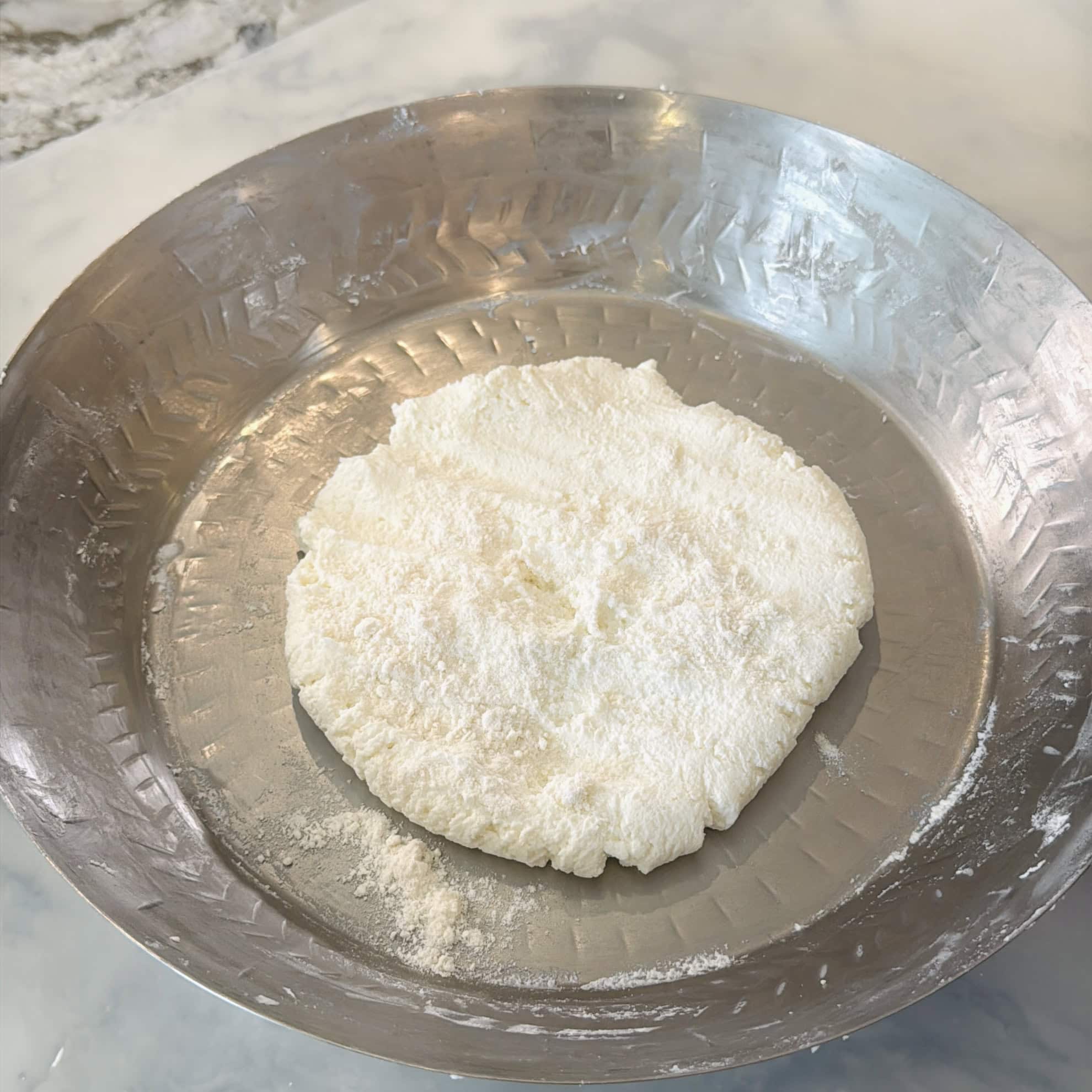 A round, flat disc of raw dough sits on a large, shallow, metal plate dusted with flour.