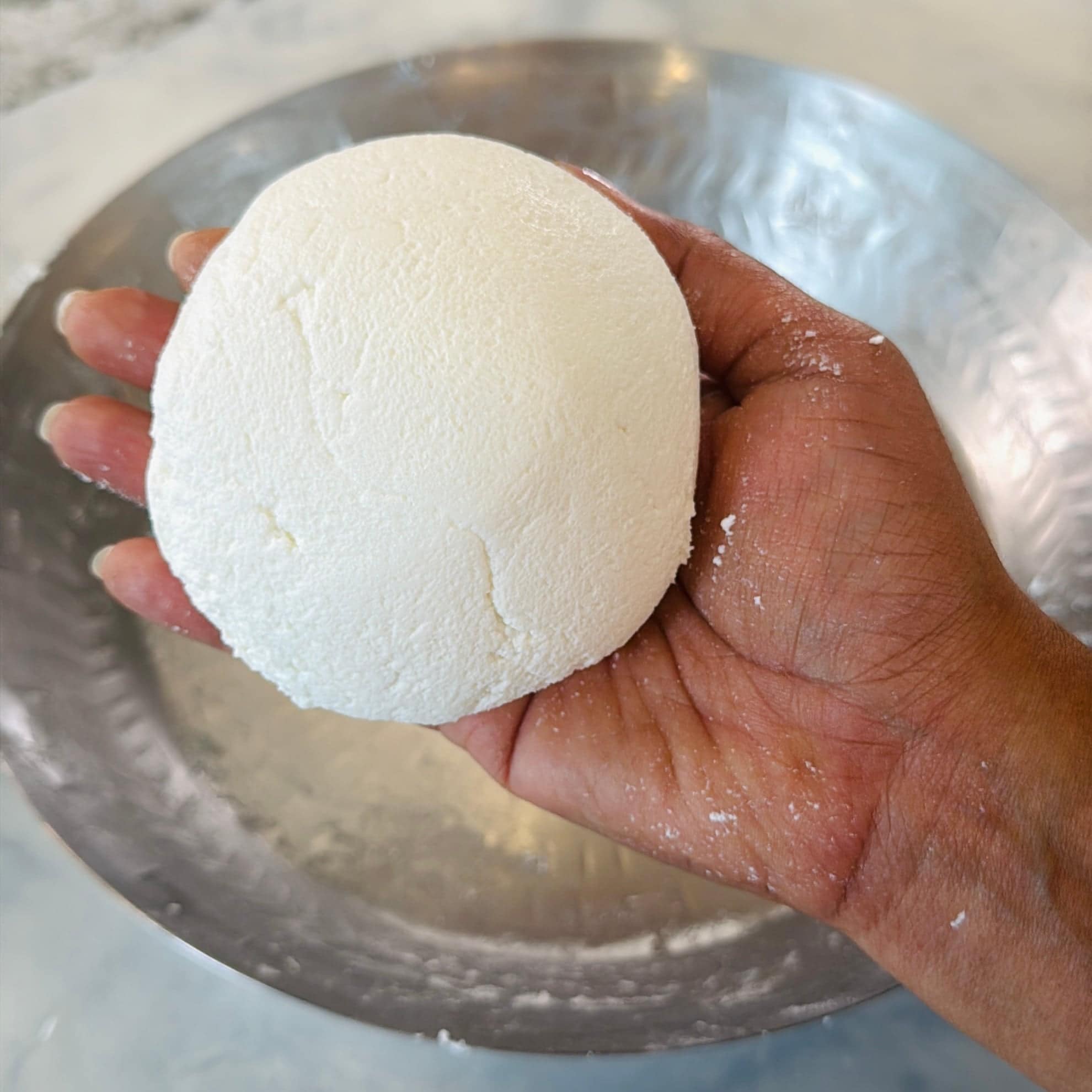 A hand holds a round, white ball of fresh paneer over a metal plate.