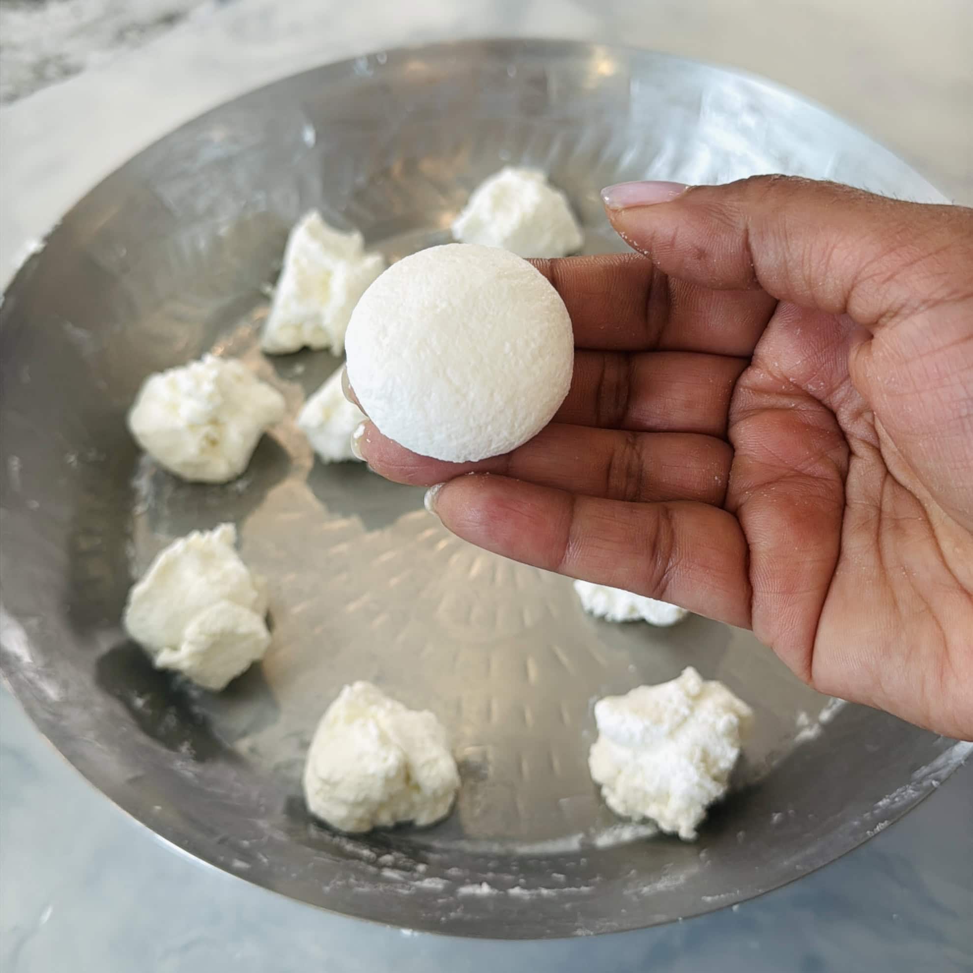 A hand holds a smooth white cheese ball over a metal plate with several unfinished cheese portions.