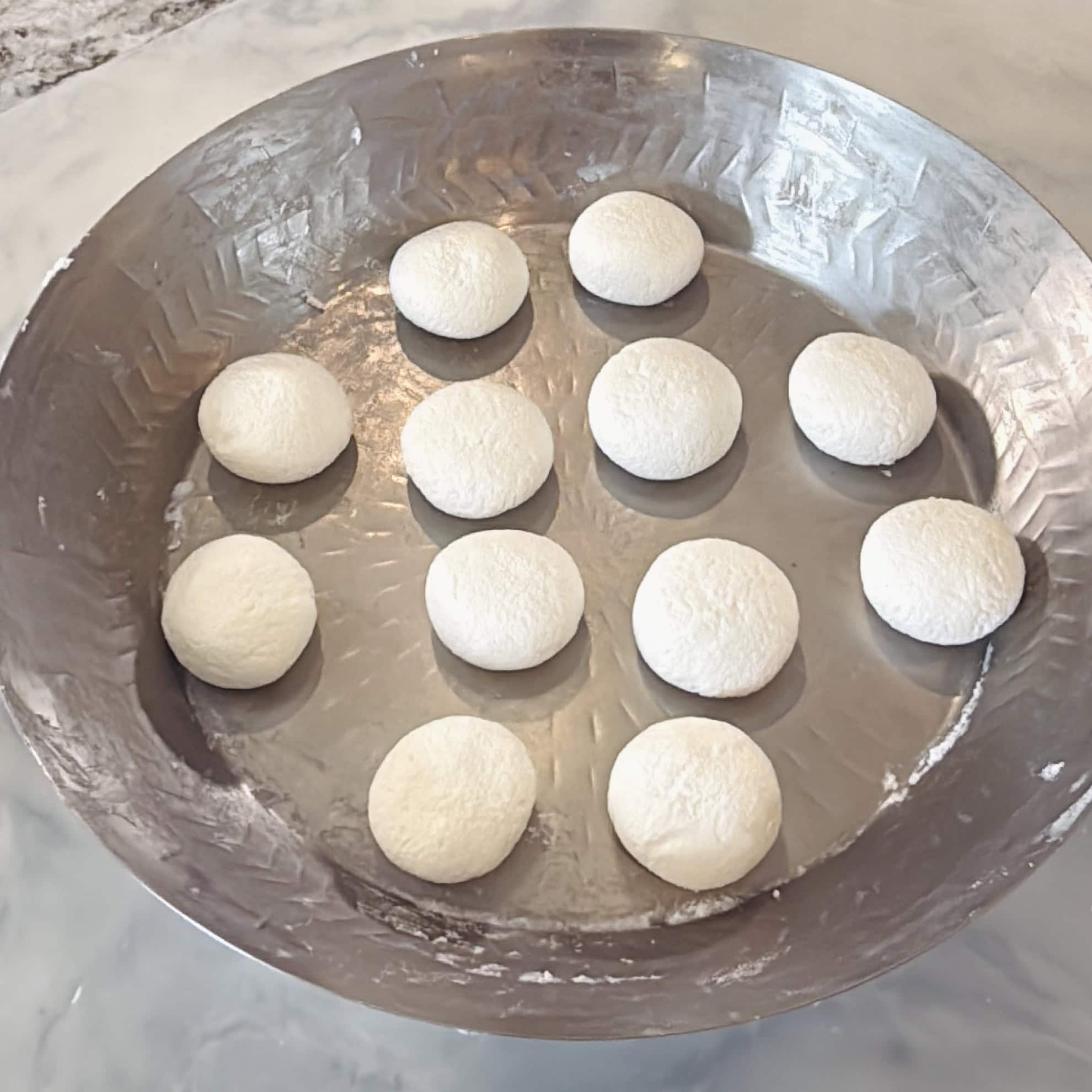 Twelve round dough balls are evenly spaced in a large, round metal tray on a marble surface.