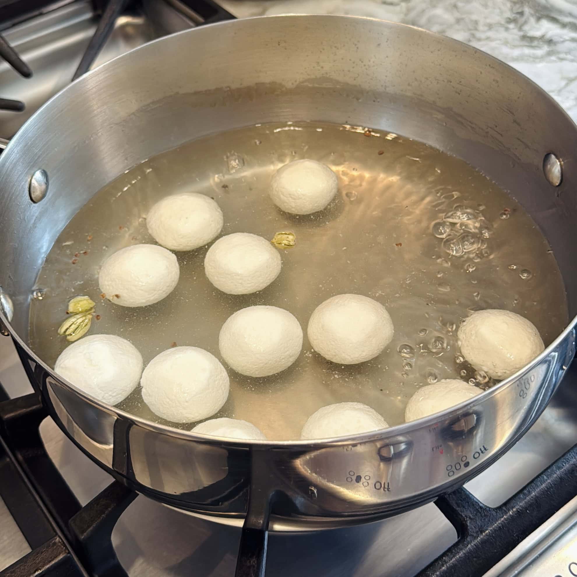 A stainless steel pot on a stove contains white balls boiling in sugar syrup, with cardamom pods visible in the liquid.