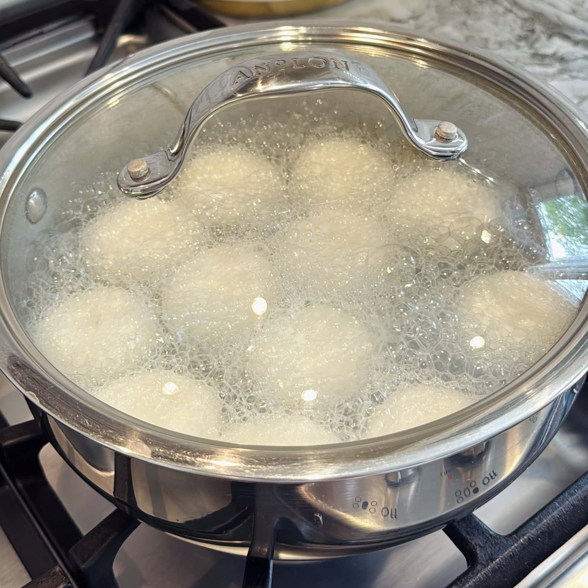 A pot of eggs is boiling on a stovetop with the lid on, creating steam and bubbles inside the pot.