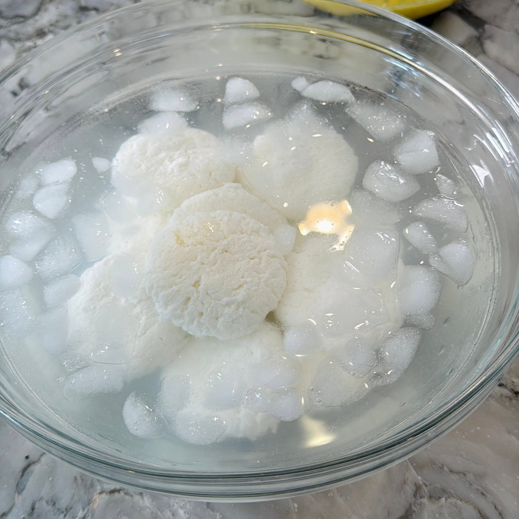 A glass bowl filled with ice water and several white, round discs of cheese submerged, on a marble countertop.