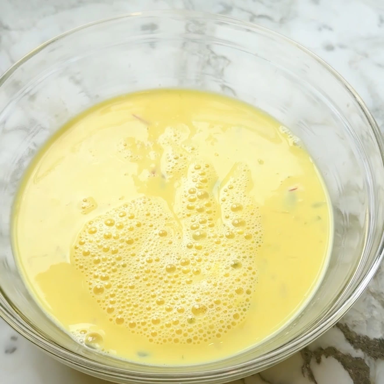 A glass bowl filled with a yellow, frothy egg mixture sits on a marble countertop.