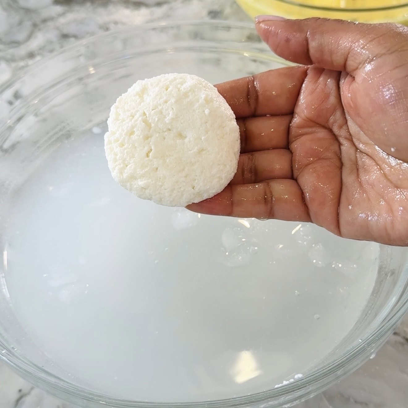 A hand holds a round, white cheese curd above a bowl of cloudy liquid, likely whey, during a cheese-making process.