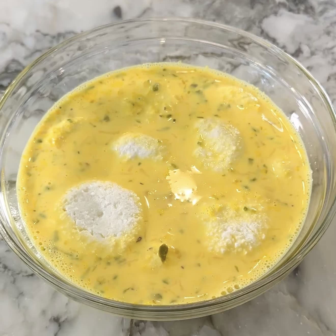 A glass bowl contains a yellow spiced yogurt mixture with four white steamed rice balls partly submerged, on a marble surface.