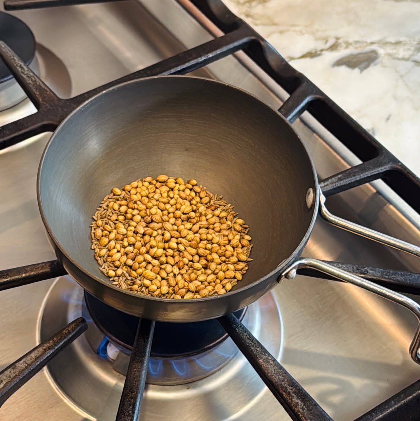 Whole coriander and cumin seeds are being dry toasted in a small metal pan over a lit gas stove burner.