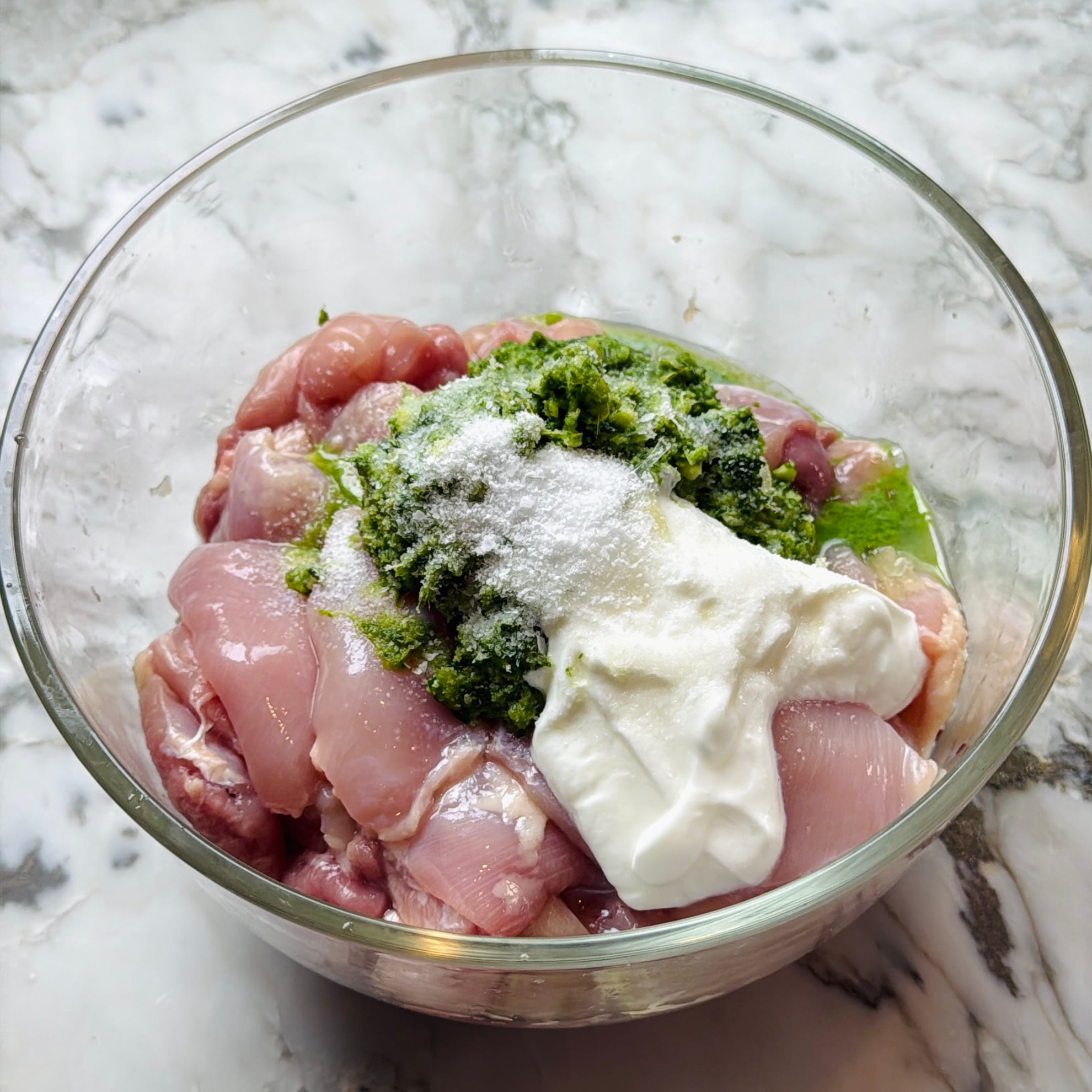 Raw chicken pieces in a glass bowl with yogurt, green herb paste, and salt on top, ready to be mixed on a marble countertop.