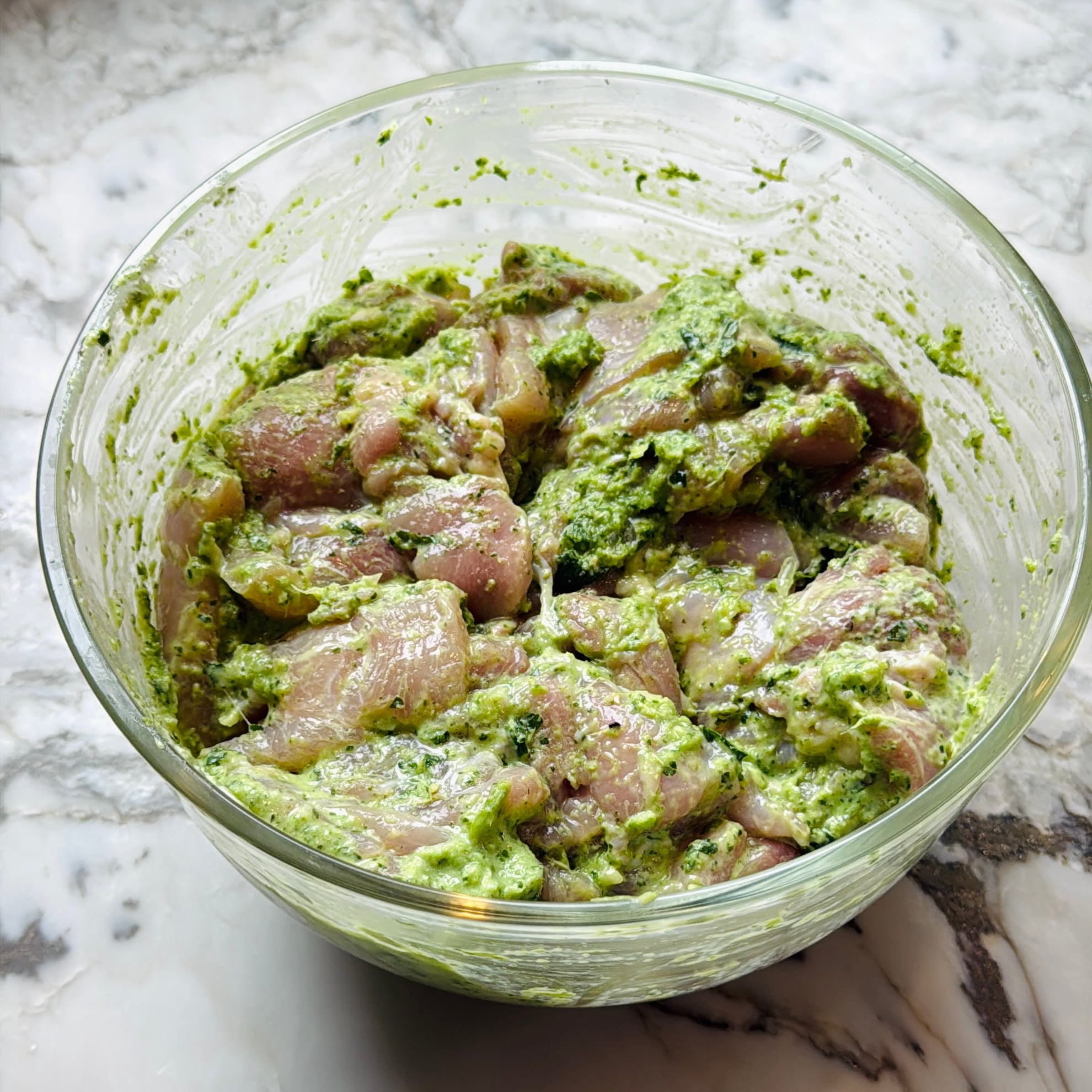Raw chicken pieces coated in a green herb marinade sit in a clear glass bowl on a marble countertop.
