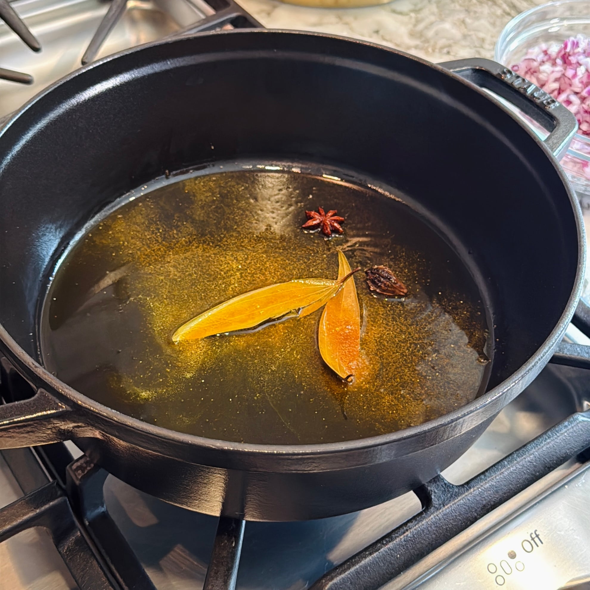 A black pot on a stove contains hot oil with bay leaves, star anise, and a black cardamom pod.