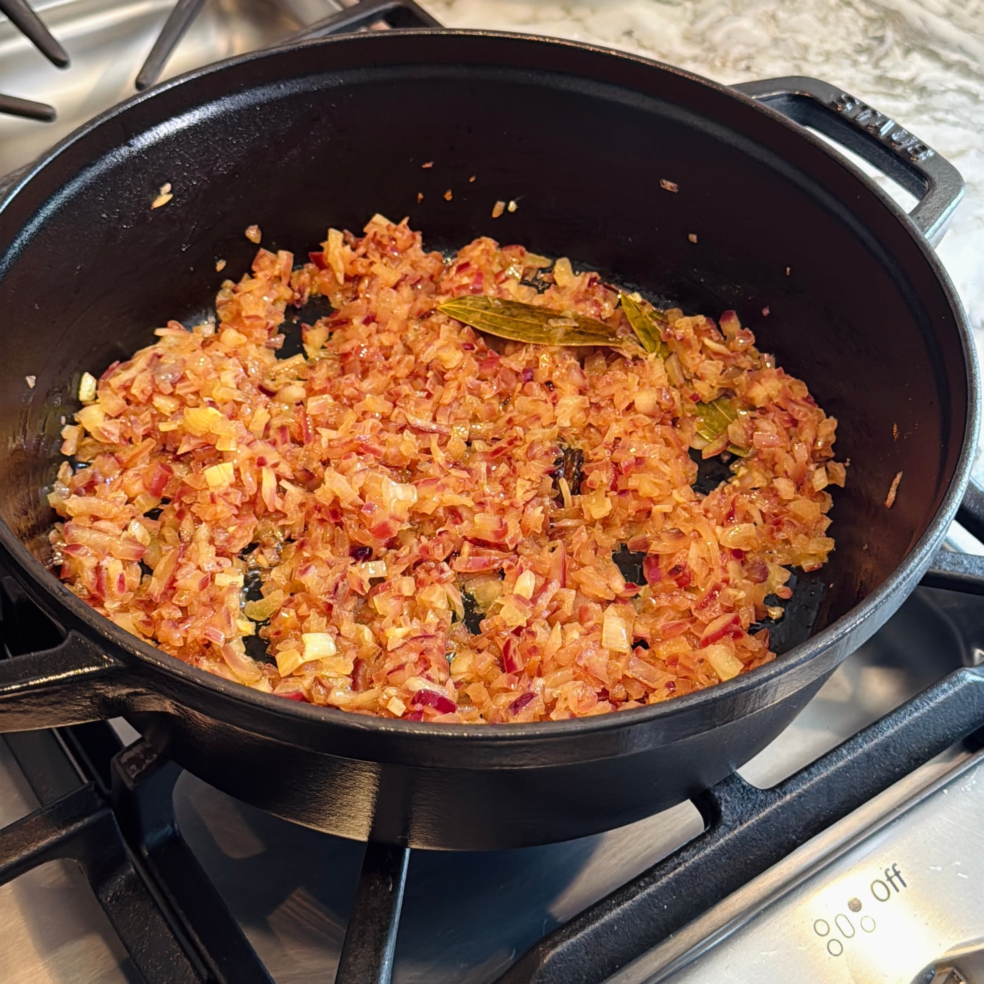 Chopped onions sautéing with spices and a bay leaf in a black pot on a stove.