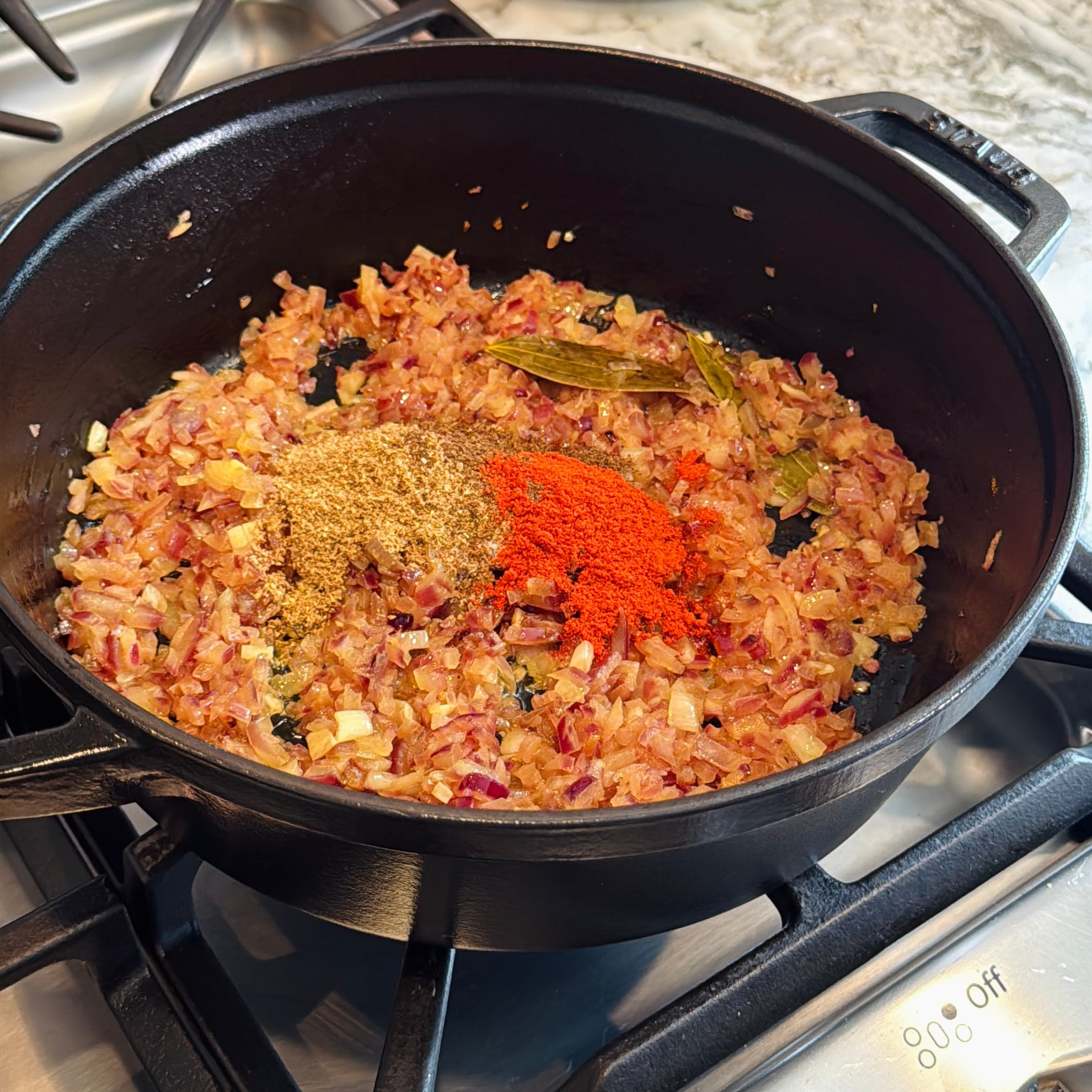 Chopped onions sautéing in a black pot on a stove with ground spices and a bay leaf added on top.