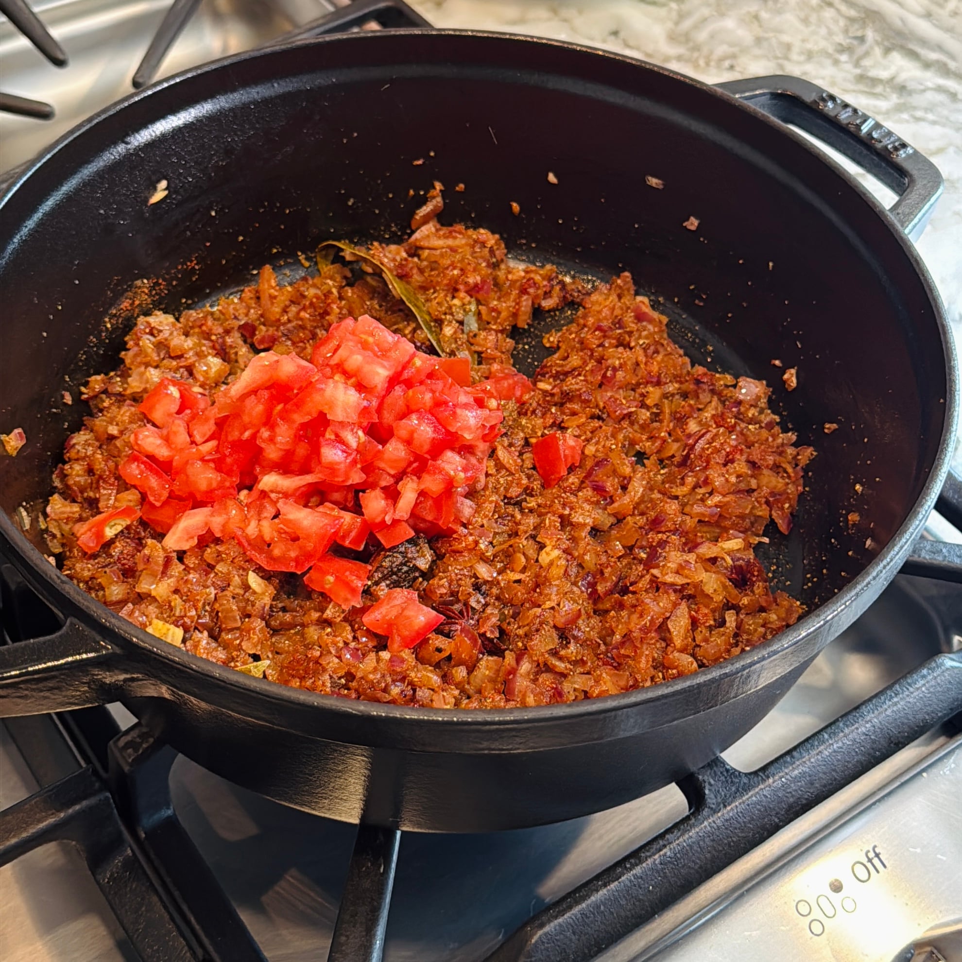 Diced tomatoes are added to a pot of sautéed onions, spices, and seasonings on a stovetop.