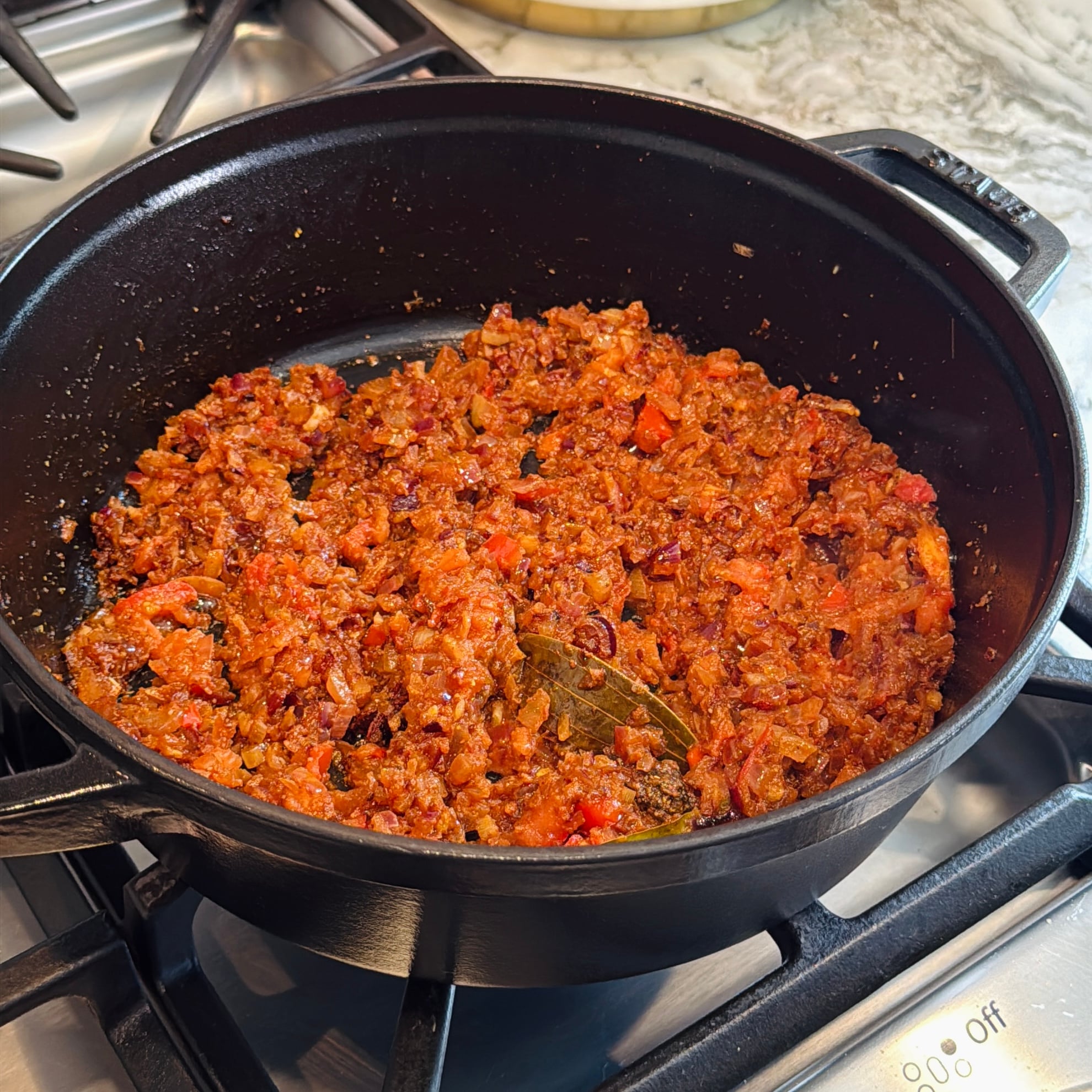 A black pot on a stove contains sautéed onions, tomatoes, and spices, including a visible bay leaf.