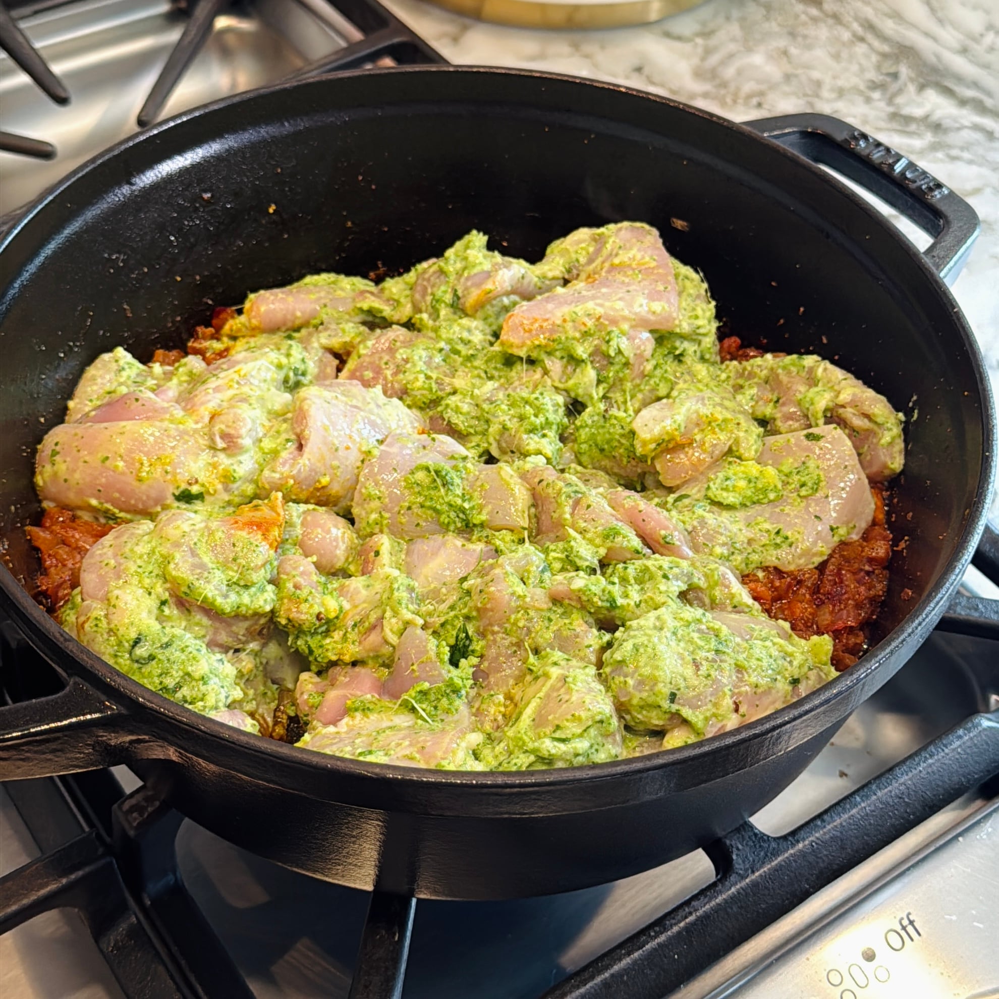 Raw chicken pieces coated with green marinade are arranged in a black cast iron pot on a stovetop, with sautéed onions or spices visible underneath.
