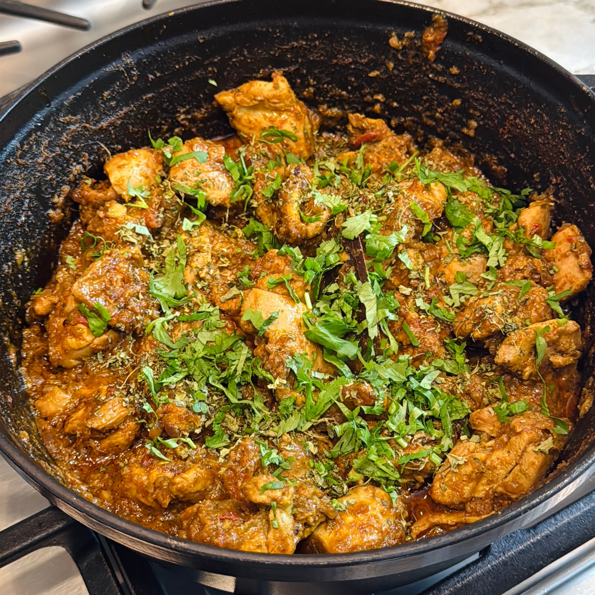 A pot of cooked chicken curry garnished with chopped fresh herbs sits on a stovetop, showing chunks of chicken in a thick, spiced sauce.