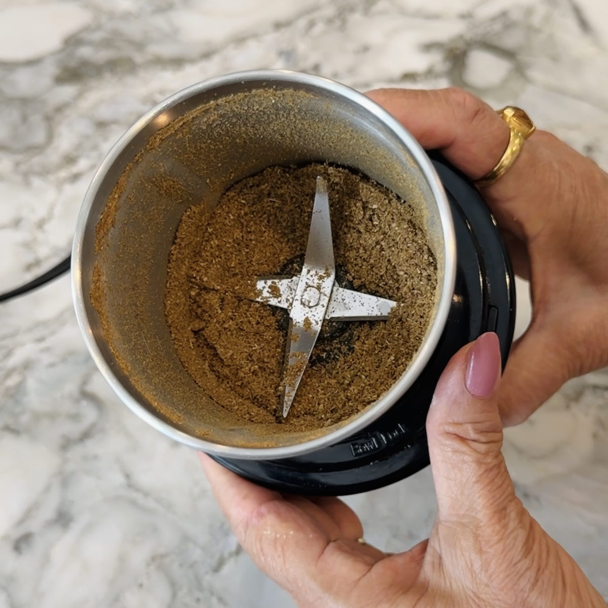 A hand holds a spice grinder with ground spices inside on a marble countertop.