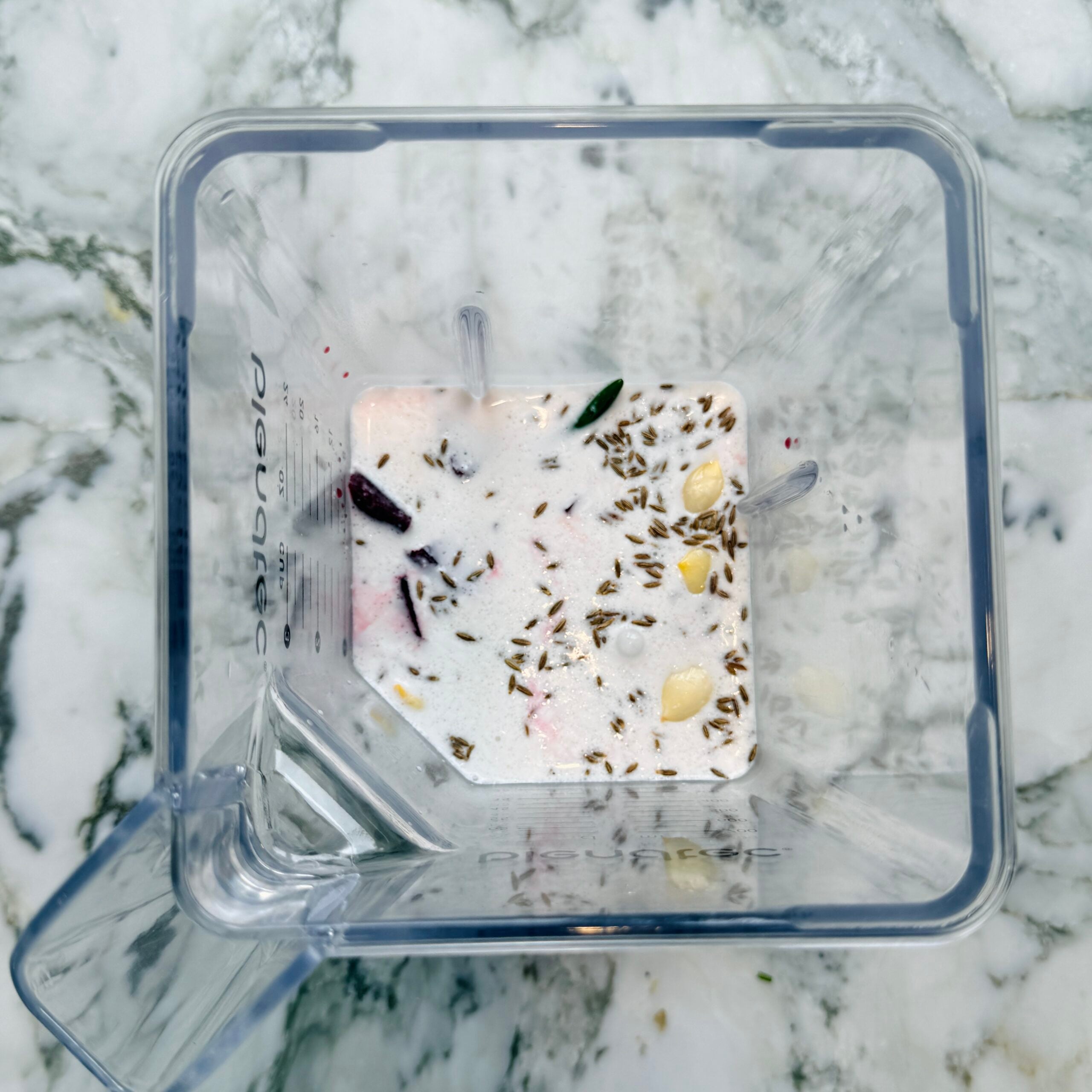 A blender jar with coconut milk and spices on a white marble countertop, viewed from above.