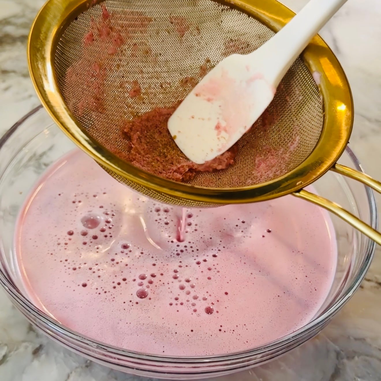 A spatula presses pink liquid through a fine mesh strainer over a clear bowl on a marble countertop.
