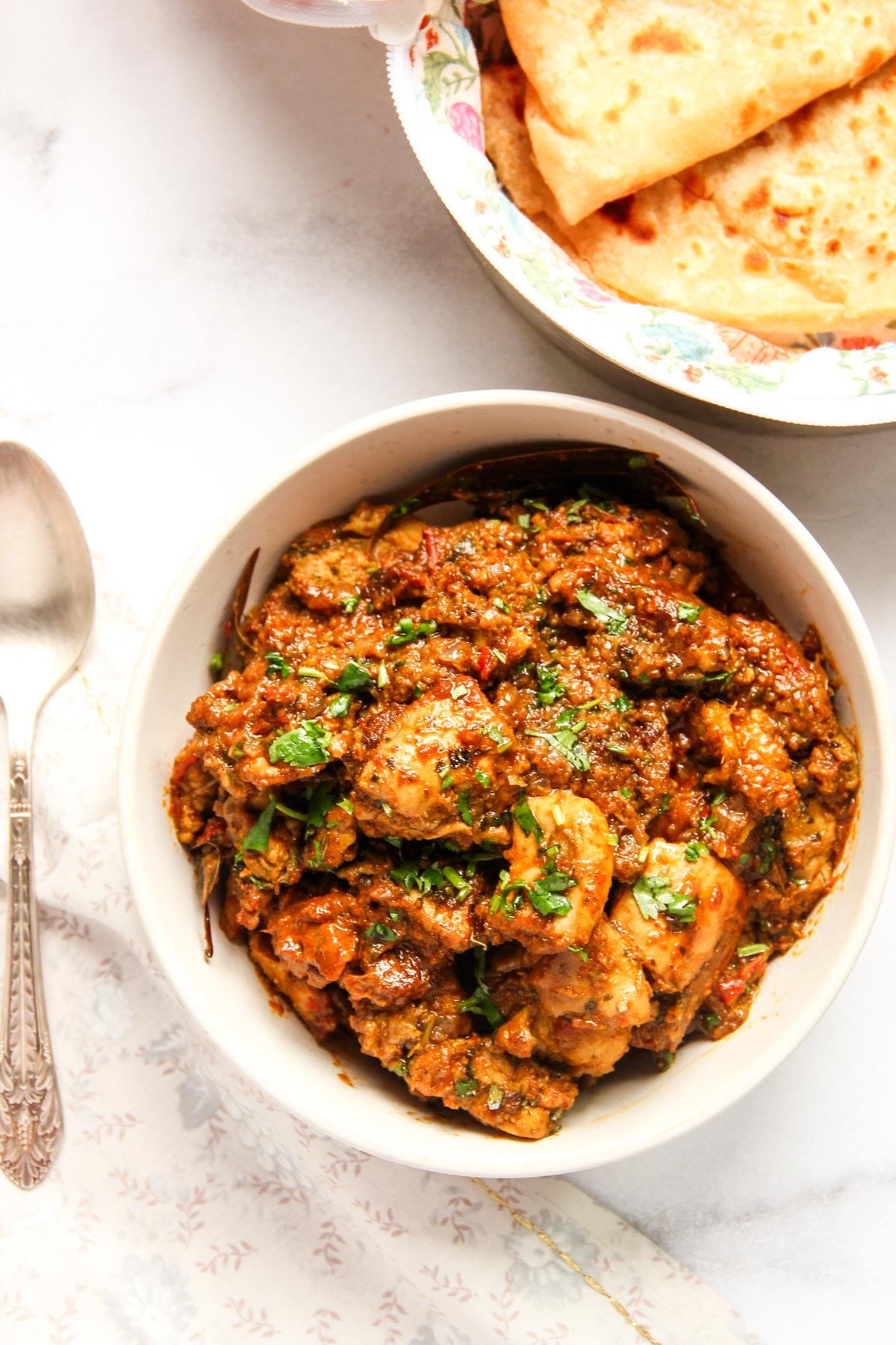 A bowl of spiced chicken curry garnished with cilantro sits next to a plate of folded flatbread, with a silver spoon on a white cloth nearby.