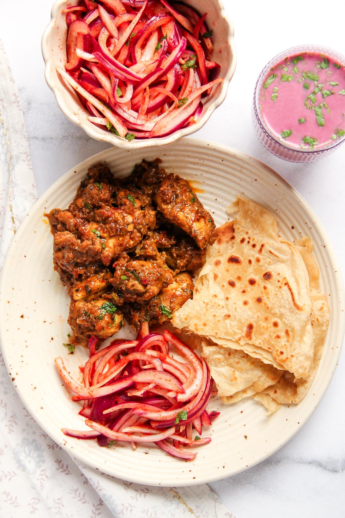 A plate with chicken sukka, chapati, and a onion salad, accompanied by a bowl of salad and a glass of solkadhi.