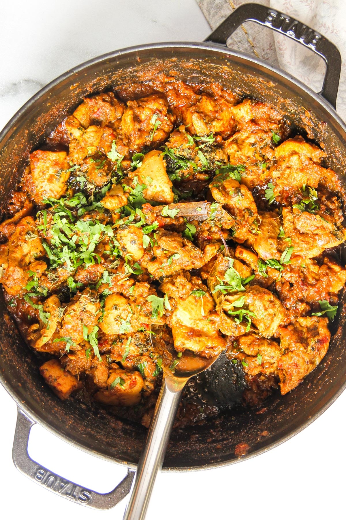 A pot of cooked chicken curry garnished with chopped cilantro, with a serving spoon resting inside the dish.