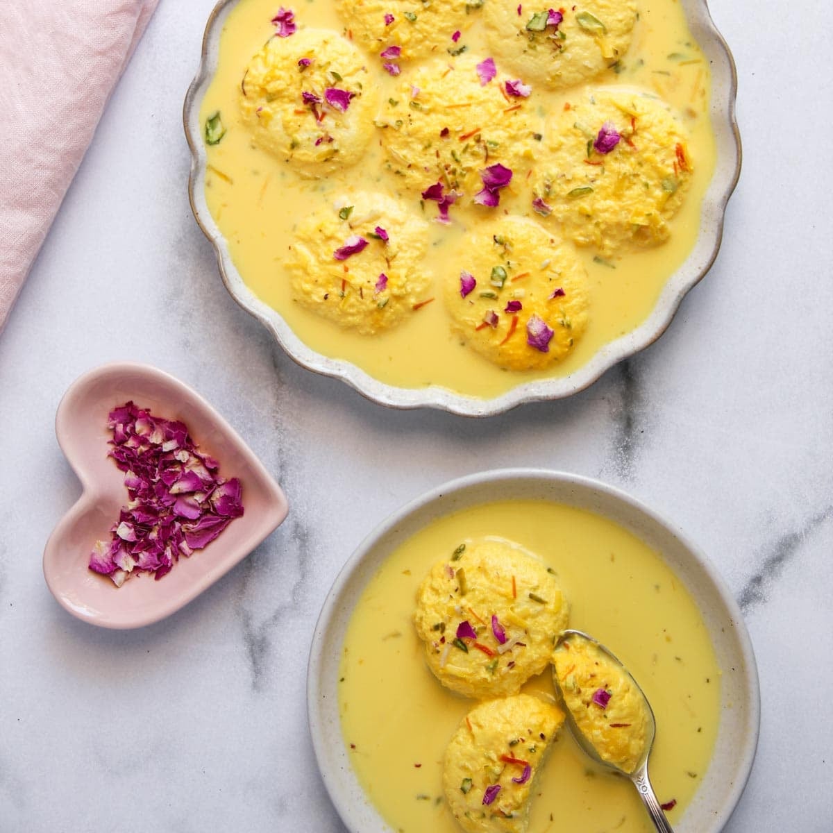 Two bowls of ras malai served in saffron milk and garnished with rose petals, with a heart-shaped dish holding dried rose petals on the side.