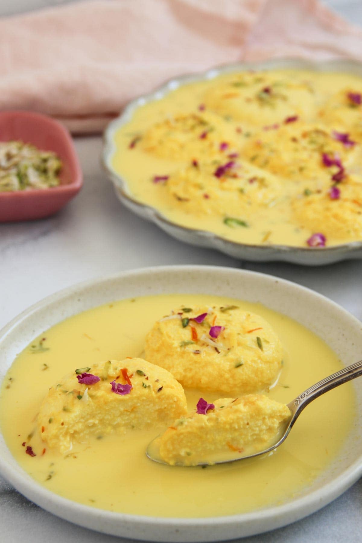 Three pieces of rasmalai in yellow saffron milk on a plate, garnished with chopped nuts and rose petals, with more rasmalai in the background.