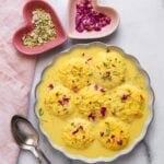 A bowl of ras malai garnished with nuts and rose petals, with two spoons and small heart-shaped dishes of sliced nuts and dried rose petals on the side.