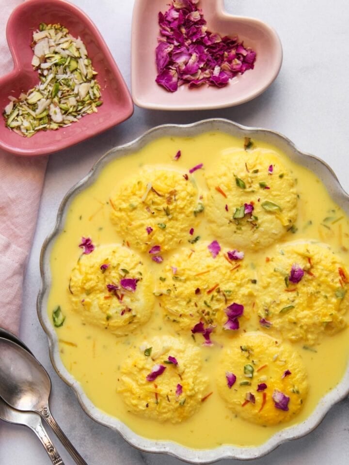 A bowl of ras malai garnished with nuts and rose petals, with two spoons and small heart-shaped dishes of sliced nuts and dried rose petals on the side.