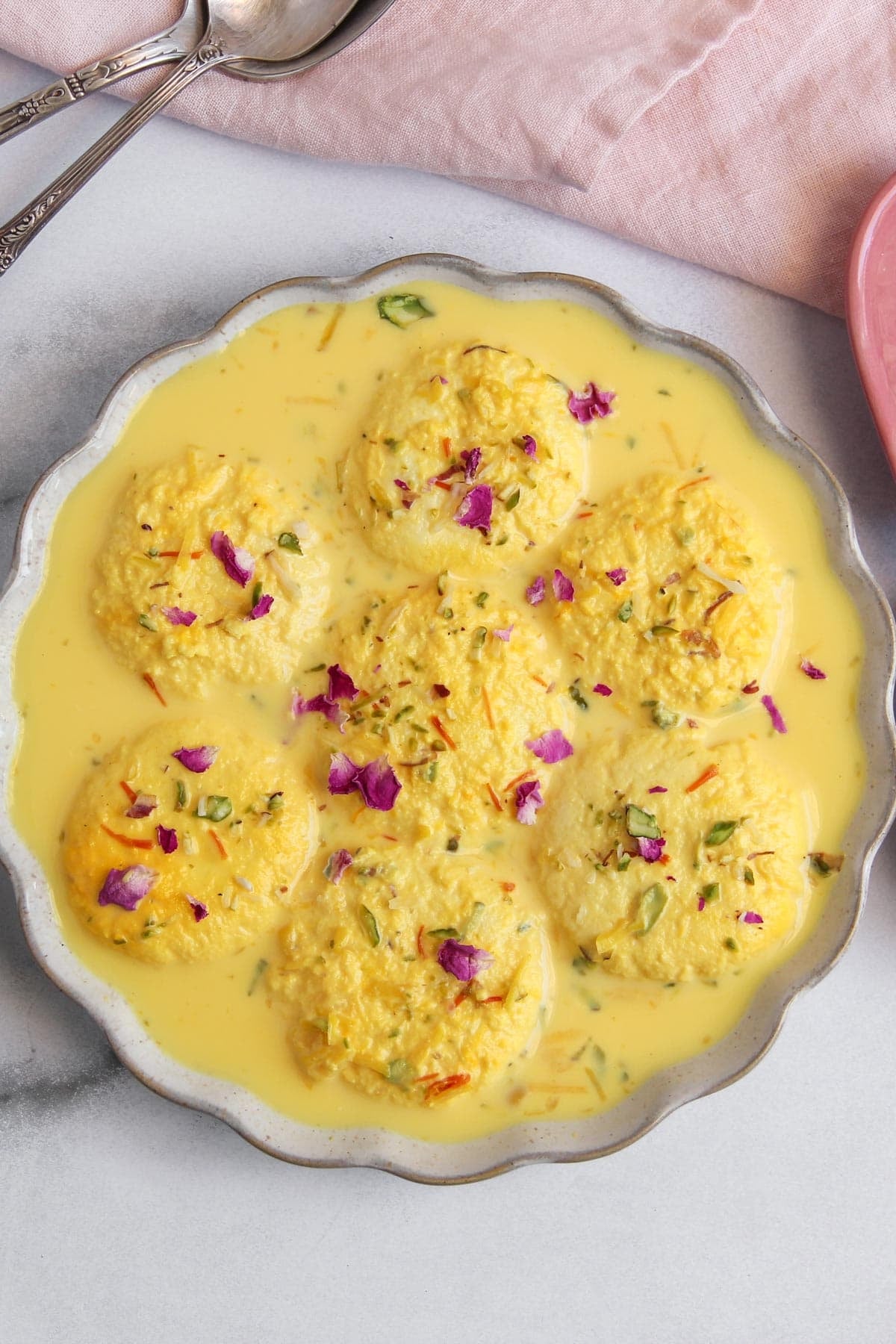 A round white dish with six yellow ras malai pieces soaked in creamy saffron milk, garnished with chopped pistachios and rose petals.