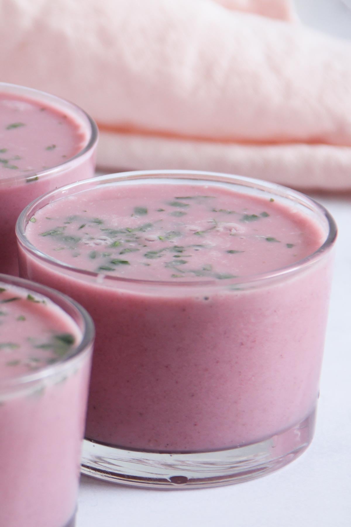 Three clear glasses filled with solkadhi garnished with cilantro, set on a white surface with a pale pink cloth in the background.