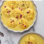 A bowl and plate of rasmalai, round yellow cheese dumplings in creamy yellow milk, garnished with rose petals and pistachios, on a light surface with two spoons nearby.