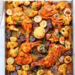 Sheet pan with roasted tandoori chicken, cauliflower, sliced potatoes, lemon slices, and herbs on a marble background.