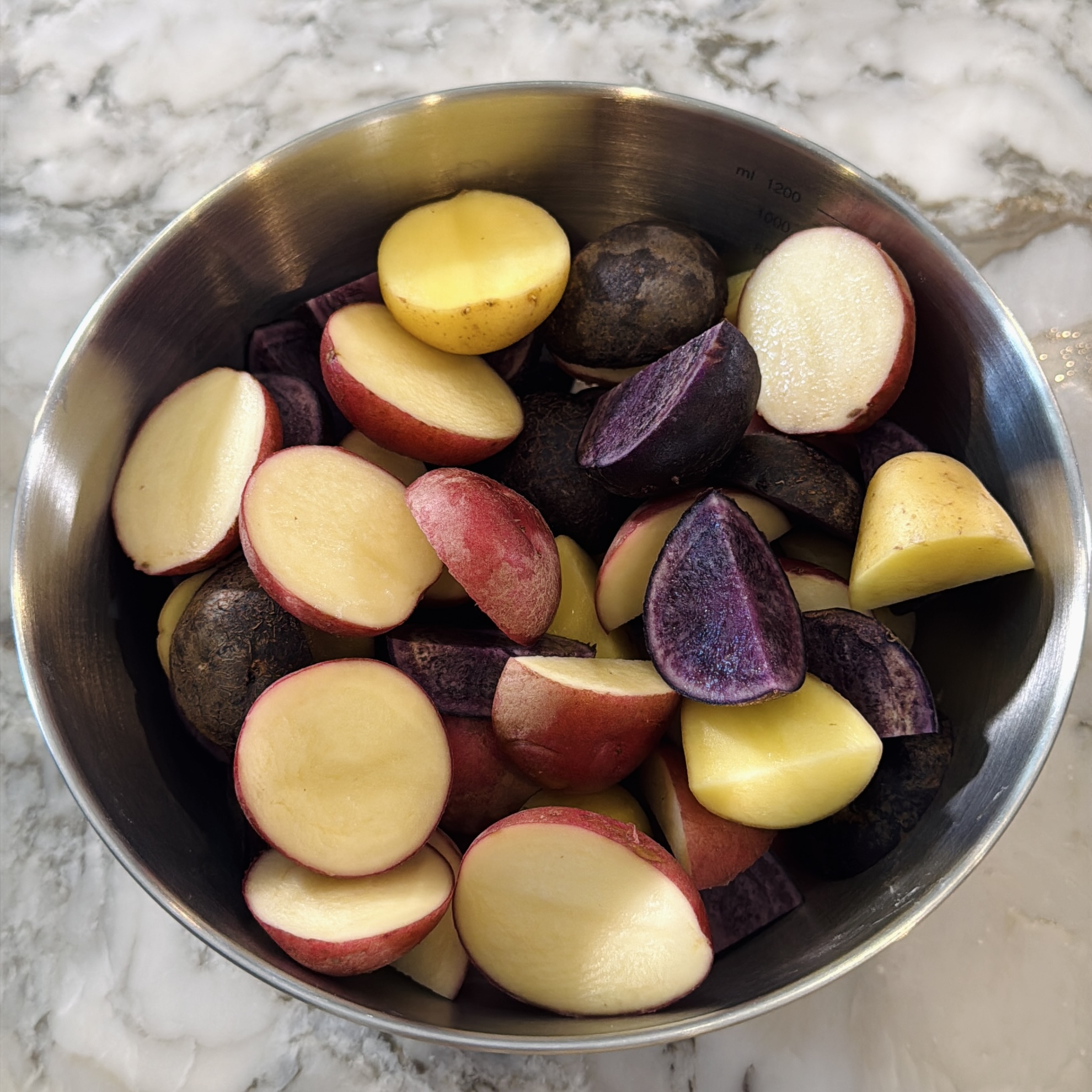 A metal bowl filled with cut pieces of red, yellow, and purple potatoes on a marble countertop.