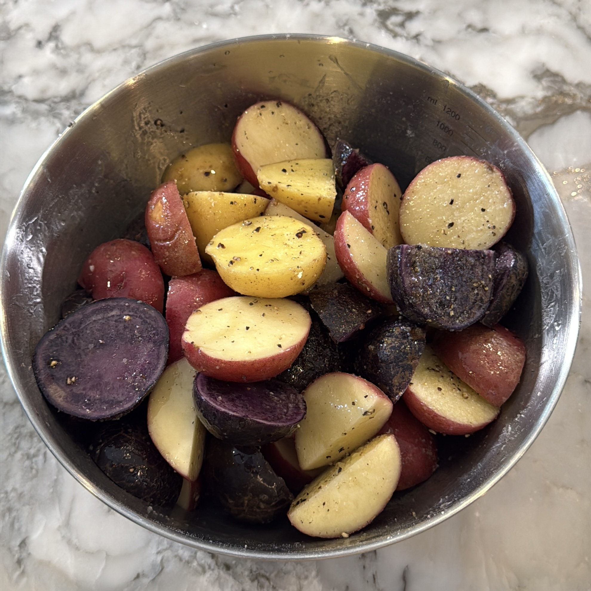 A metal bowl filled with halved red, yellow, and purple potatoes, seasoned with black pepper, sits on a marble countertop.