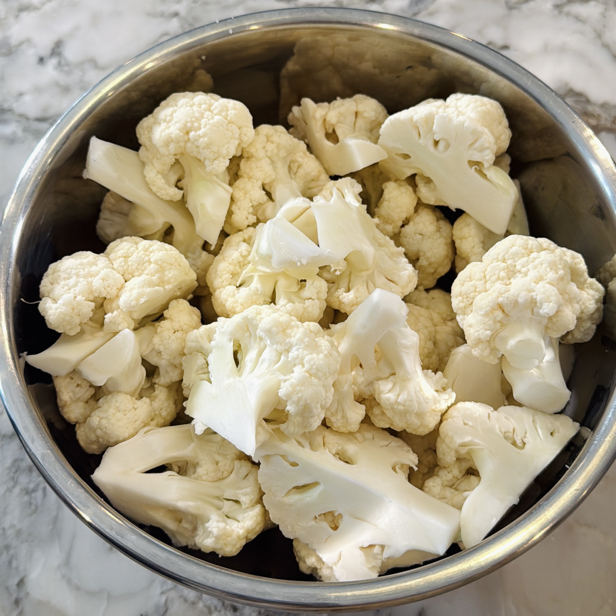 A stainless steel bowl filled with raw cauliflower florets on a marble countertop.