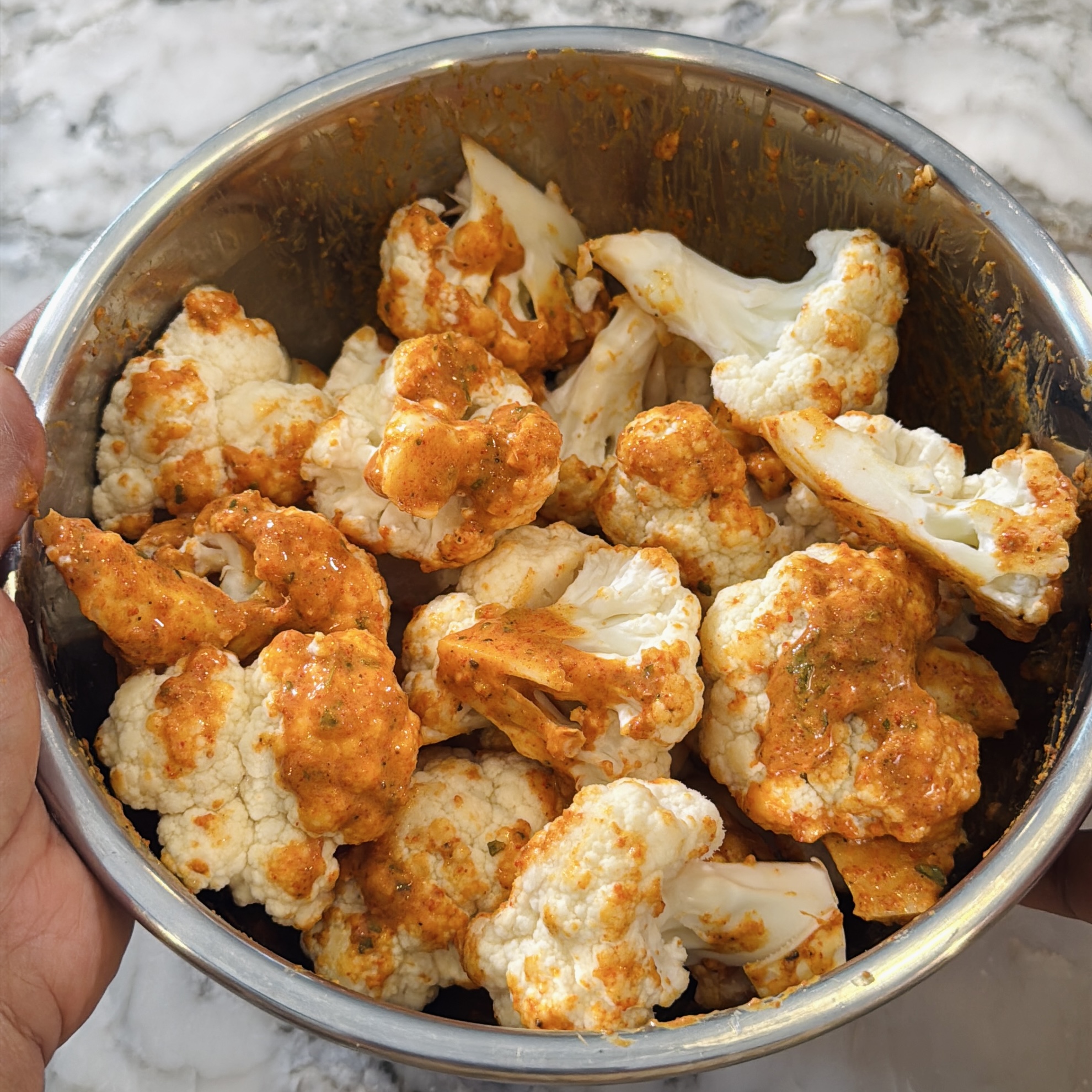 A stainless steel bowl filled with cauliflower florets coated in an orange marinade held over a marble countertop.