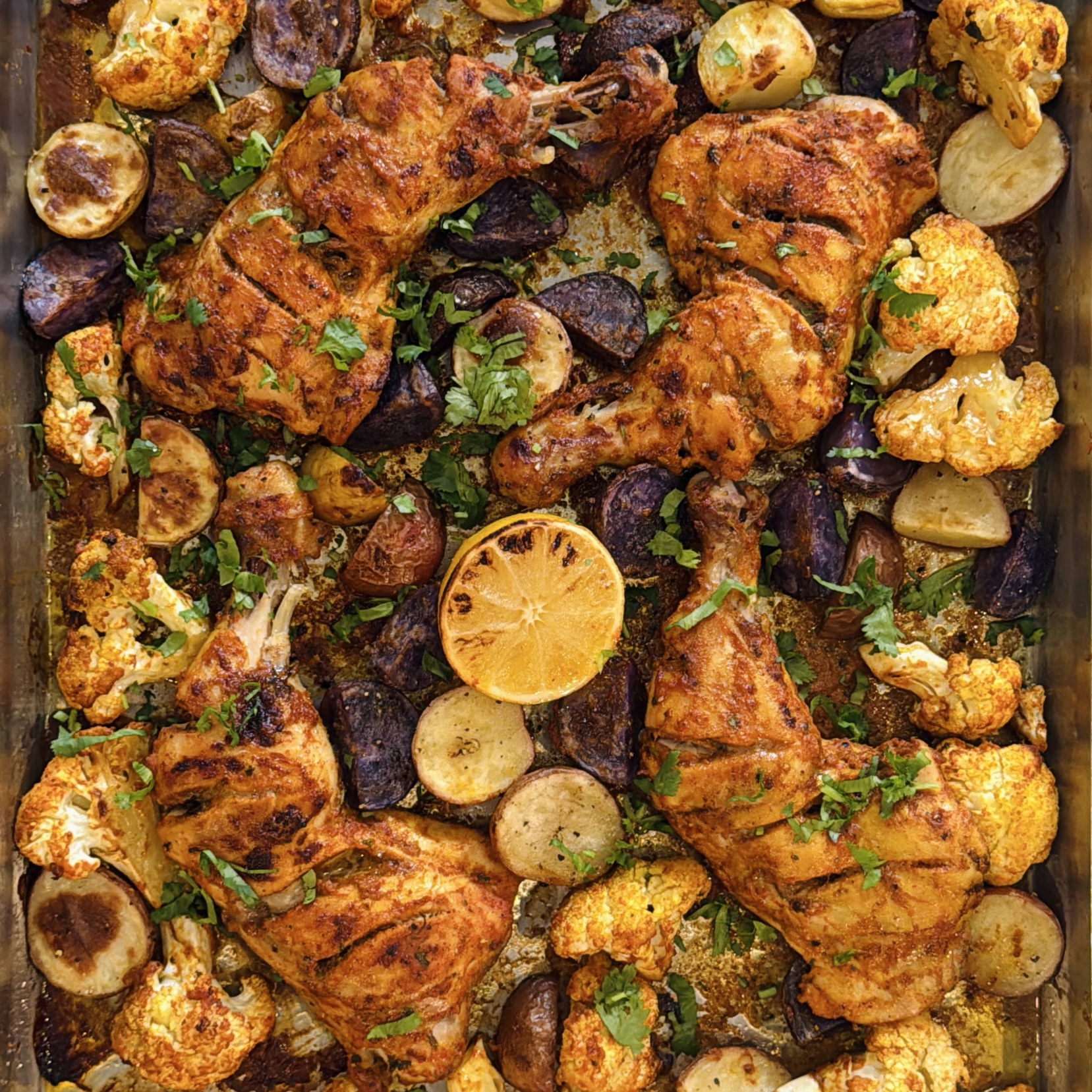 Baked chicken legs with roasted potatoes, cauliflower, and lemon slices on a sheet pan, garnished with chopped herbs.