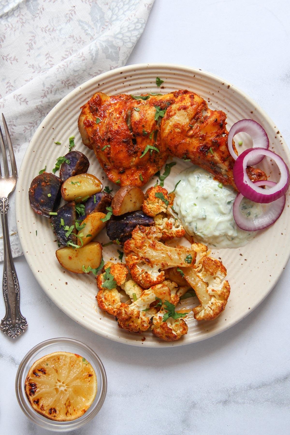 Plate with roasted chicken, sliced potatoes, cauliflower, red onion rings, and a dollop of white sauce, garnished with herbs. Grilled lemon slice and utensils are next to the plate.