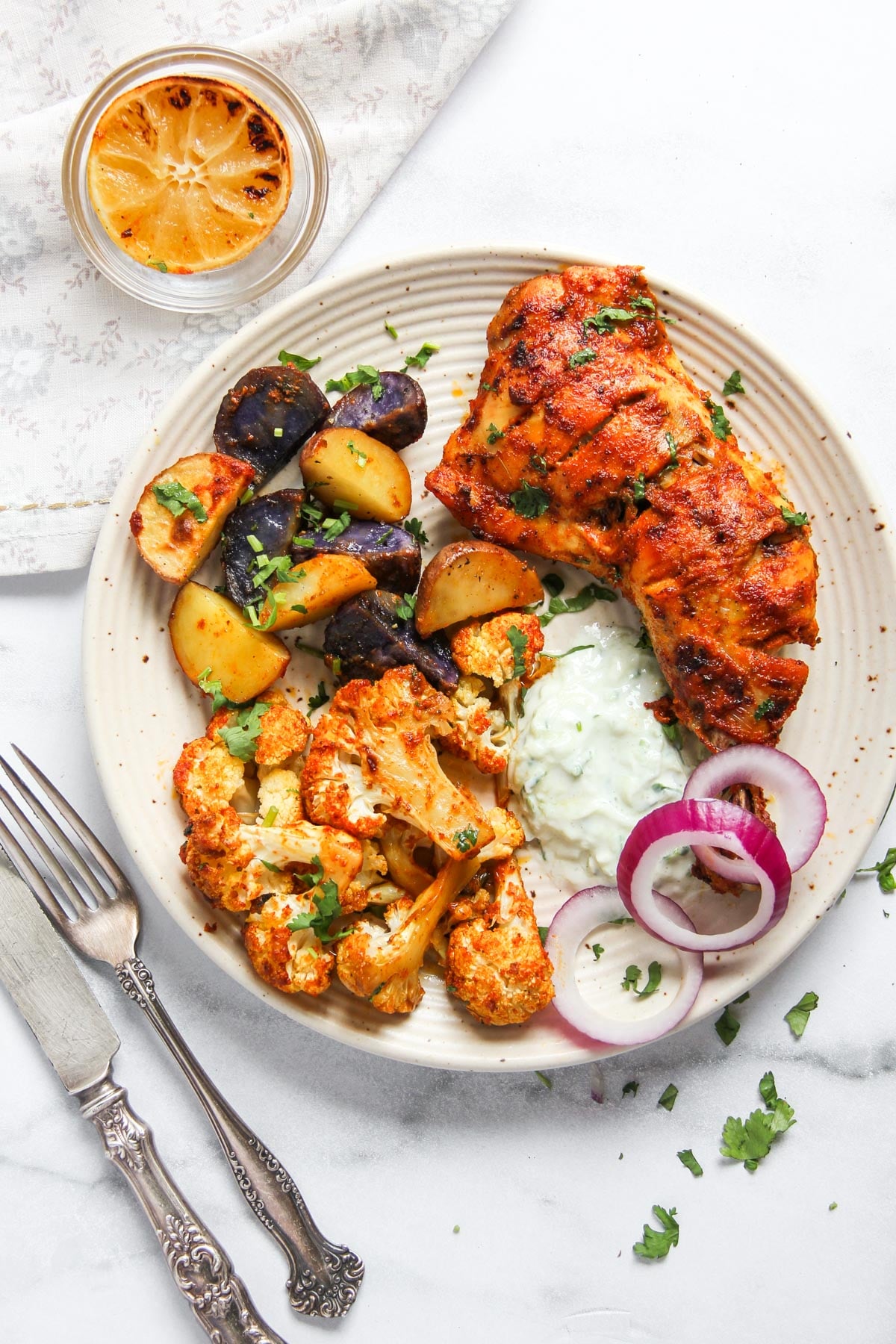 A plate with grilled chicken, roasted potatoes, roasted cauliflower, sliced red onions, a dollop of tzatziki sauce, and a drink with a slice of lemon. Knife and fork rest beside the plate.