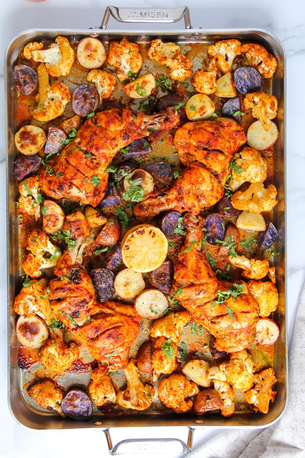 Sheet pan with roasted chicken, cauliflower, purple potatoes, onions, and lemon slices, garnished with herbs.