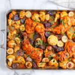 A sheet pan with roasted chicken legs, cauliflower florets, sliced purple and yellow potatoes, and halved lemons, garnished with chopped herbs.