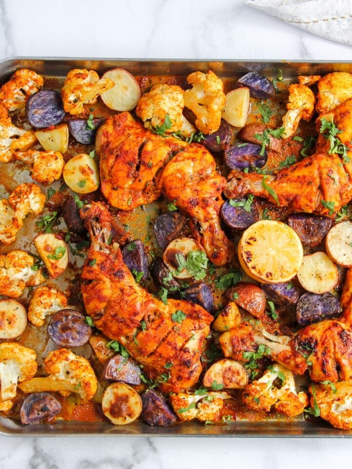 A sheet pan with roasted chicken legs, cauliflower florets, sliced purple and yellow potatoes, and halved lemons, garnished with chopped herbs.