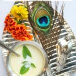 A silver tray with a bowl of panchamrit with holy basil leaves, marigold flowers, a peacock feather, and a small silver figure, on a dotted cloth.