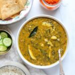 A bowl of Alu Chi Bhaji curry with colocasia leaves, served with rice, flatbread, sliced cucumbers, and a spice garnish.