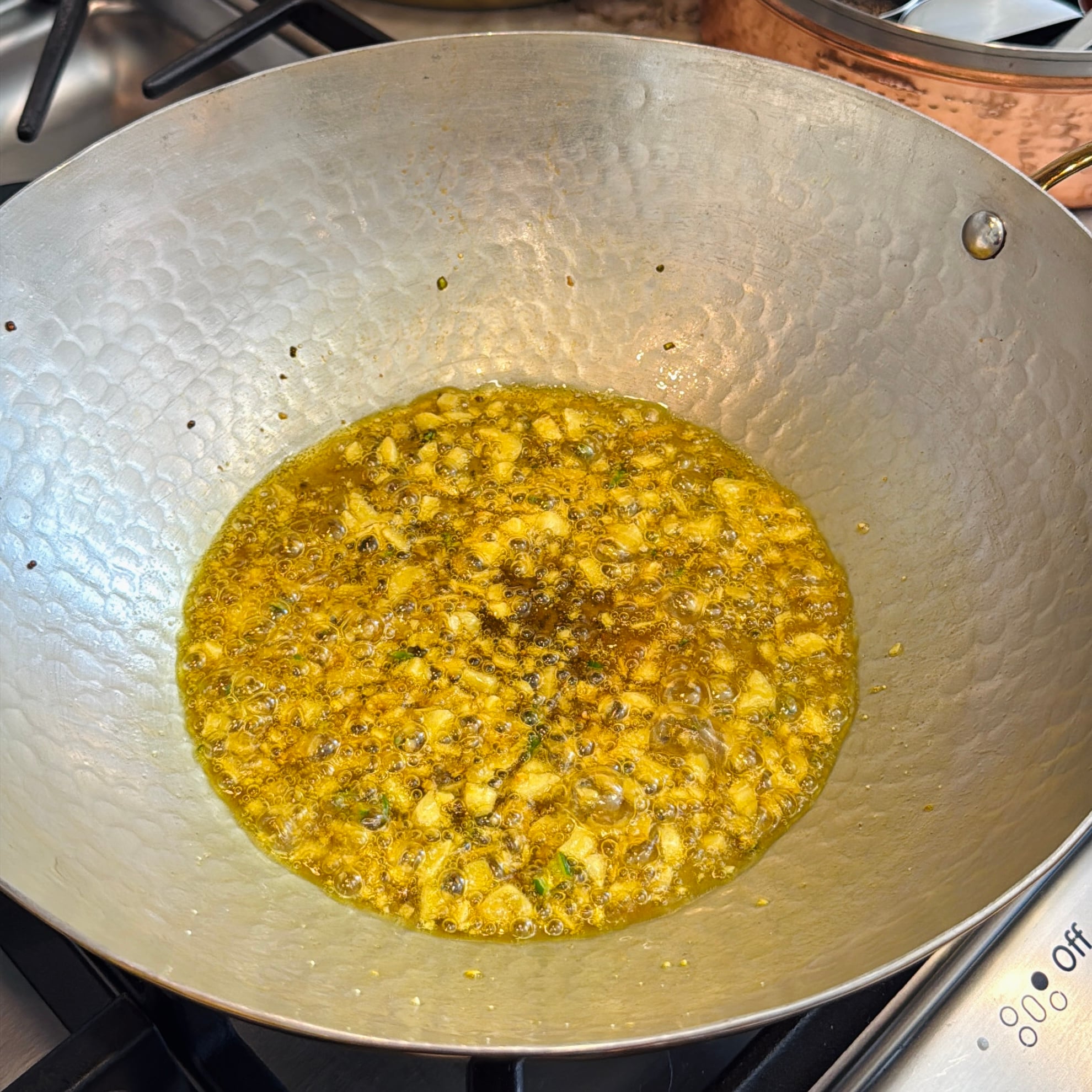 A kadhai on a stove contains a simmering tadka of mustard seeds, turmeric and garlic.